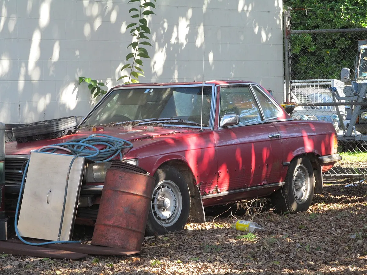 A red Mercedes-Benz SL parked on grass behind a garage, with rust bubbling through the front wing and sill